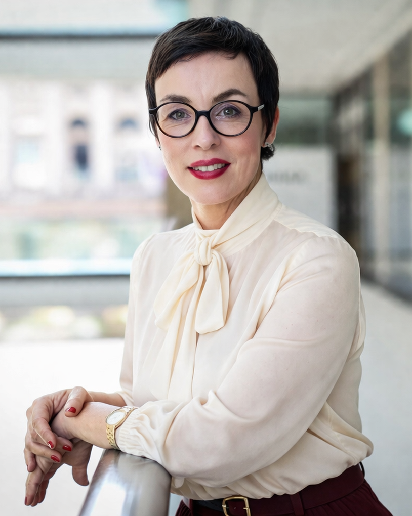Katja Fesselman von schräg vorne an einem Geländer lehnend. Schwarze Brille, kurze Haare und kurzärmliches Shirt