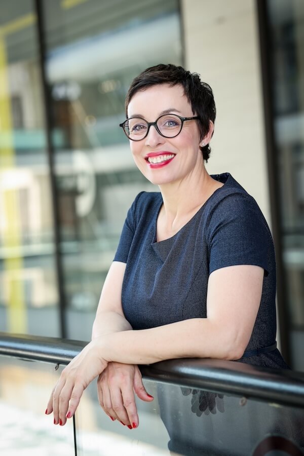 Katja Fesselman von schräg vorne an einem Geländer lehnend. Schwarze Brille, kurze Haare und kurzärmliches Shirt